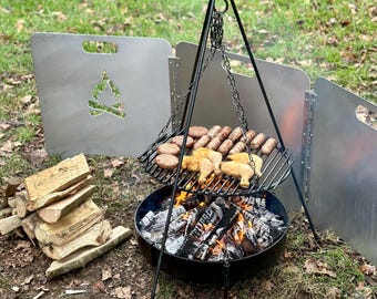 Puede incluir: Una parrilla trípode negra con una rejilla de cocción está suspendida sobre un pozo de fuego. Se están asando salchichas y trozos de pollo. Dos protectores contra el viento plateados con un recorte de fogata están a cada lado. Una pila de leña está a la izquierda.