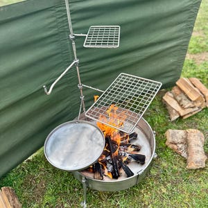 May include: A stainless steel campfire cooking setup with a fire pit, grill grates, and a cooking surface. The fire pit contains burning wood. A green windbreak is in the background. Firewood is stacked nearby.