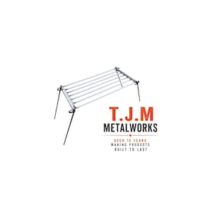 May include: A rectangular metal camping grill grate with four thin, angled legs. The text T.J.M METALWORKS is printed in orange and black, followed by the text OVER 10 YEARS MAKING PRODUCTS BUILT TO LAST, positioned below the metal grill structure.