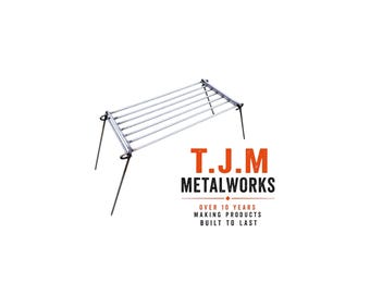 May include: A rectangular metal camping grill grate with four thin, angled legs. The text T.J.M METALWORKS is printed in orange and black, followed by the text OVER 10 YEARS MAKING PRODUCTS BUILT TO LAST, positioned below the metal grill structure.