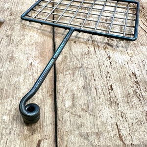 May include: A handcrafted metal grilling basket with a rectangular grid and a long handle ending in a curled design. The basket is made of dark metal and is designed for grilling food. The handle is attached to the center of the basket.