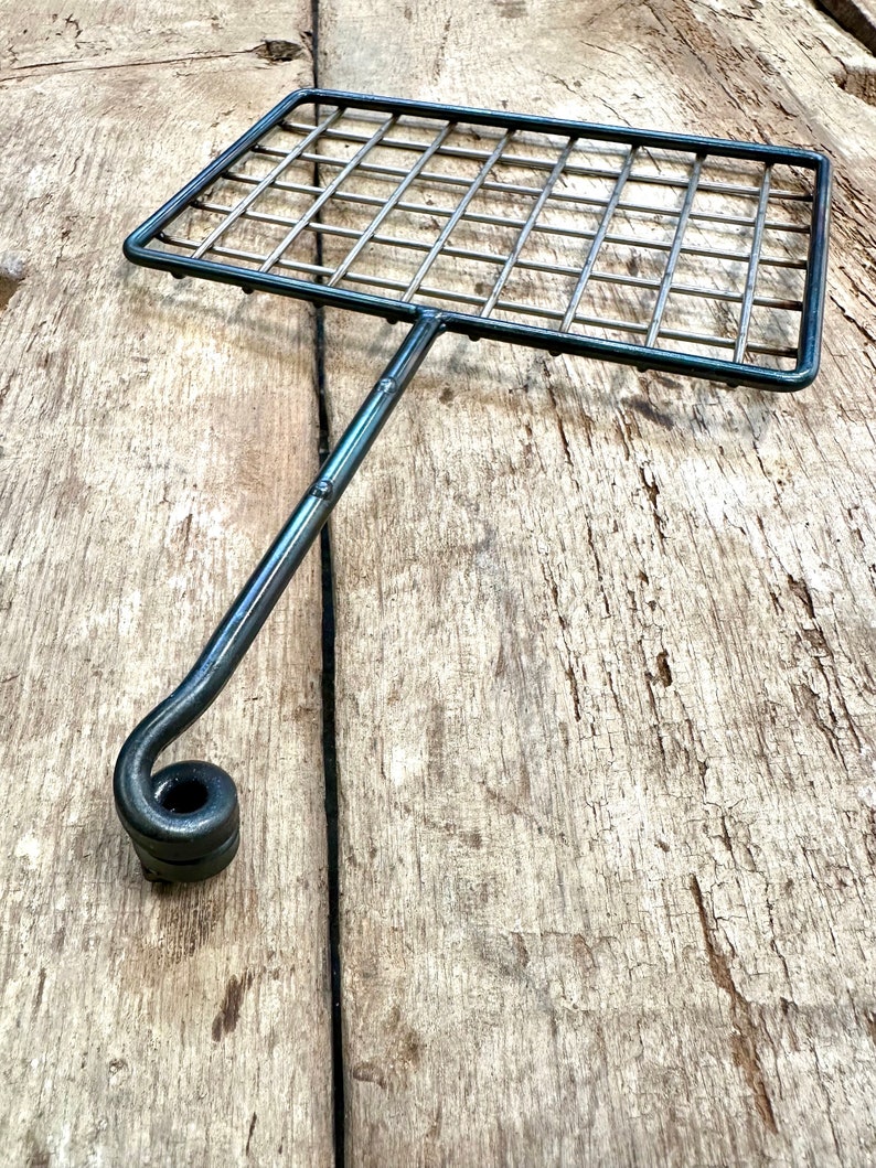 May include: A handcrafted metal grilling basket with a rectangular grid and a long handle ending in a curled design. The basket is made of dark metal and is designed for grilling food. The handle is attached to the center of the basket.
