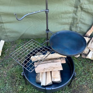 May include: A black metal fire pit with a grill grate, a round cooking surface, and a hanging hook. The fire pit is sitting on a green grassy surface.