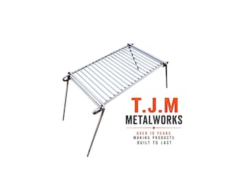 May include: A rectangular silver metal camping grill grate with four thin, foldable legs. The text T.J.M METALWORKS OVER 10 YEARS MAKING PRODUCTS BUILT TO LAST is printed in orange and black next to the grill. The grill features parallel metal bars.