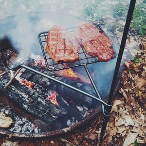May include: Two steaks grilling on a metal grate over a campfire. The steaks are reddish-brown and have grill marks. The fire is burning with visible flames and smoke. The grill is supported by a black metal stand.