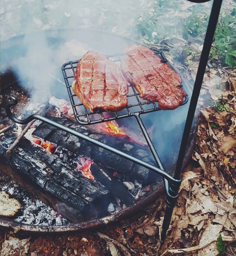 May include: Two steaks grilling on a metal grate over a campfire. The steaks are reddish-brown and have grill marks. The fire is burning with visible flames and smoke. The grill is supported by a black metal stand.