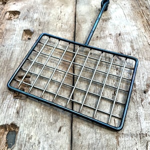 May include: A black metal grilling basket with a rectangular grid design and a long handle. The basket is made of thin metal rods and is designed for grilling food. The handle has a loop at the end for hanging.