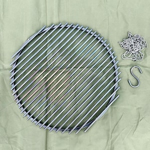 Könnte beinhalten: Ein runder Grillrost aus Metall mit einer Kette und einem Haken. Der Rost besteht aus dünnen Metallstäben, die miteinander verschweißt sind, um einen Kreis zu bilden. Die Kette ist mit einem Haken am Rost befestigt.