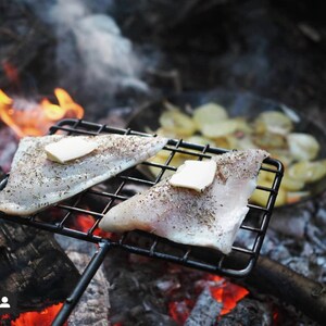 May include: Two seasoned fish fillets with pats of butter on a black metal grill over a campfire. Sliced potatoes are visible in a pan in the background. The scene evokes outdoor cooking and a rustic meal.