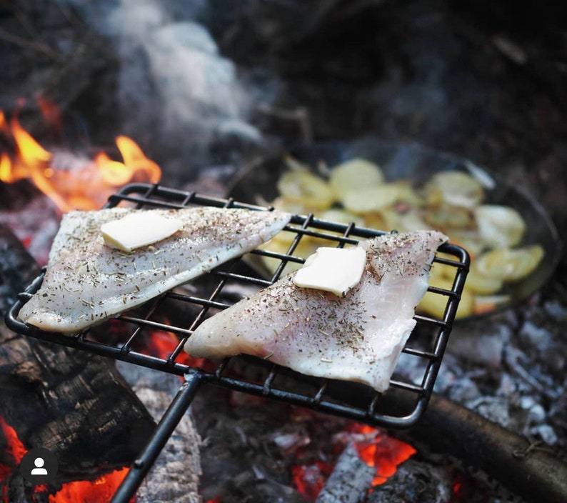 May include: Two seasoned fish fillets with pats of butter on a black metal grill over a campfire. Sliced potatoes are visible in a pan in the background. The scene evokes outdoor cooking and a rustic meal.