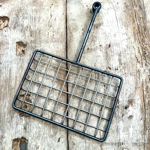 May include: A rectangular metal grilling basket with a long handle and a grid pattern. The basket is made of silver-colored metal and has a dark, possibly black, frame. The handle has a loop at the end. The basket is shown on a weathered wooden surface.