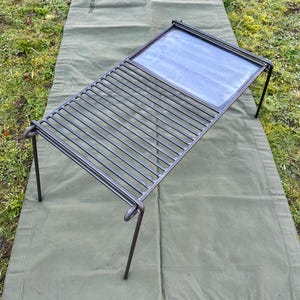 May include: A black metal grill with a flat cooking surface and a grate. The grill is on a green canvas tarp.