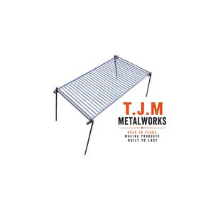 May include: A rectangular metal campfire grill grate with four foldable legs. The text T.J.M METALWORKS OVER 10 YEARS MAKING PRODUCTS BUILT TO LAST is printed in orange and black next to the grill. The grate features a series of parallel metal bars.