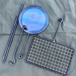 Puede incluir: Un juego de utensilios de cocina para acampar, que incluye una sartén redonda de metal, una parrilla cuadrada de metal y tres varillas de metal. Los utensilios de cocina están dispuestos sobre una lona verde.