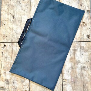 Pode incluir: Uma bolsa tote de lona azul escura com al&ccedil;as pretas. A bolsa est&aacute; deitada sobre uma superf&iacute;cie de madeira.
