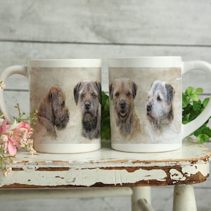 May include: Two white ceramic mugs featuring images of Border Terriers. The mugs display detailed portraits of the dogs in different poses, set against a neutral, textured backdrop. The mugs are on a distressed white wooden surface.