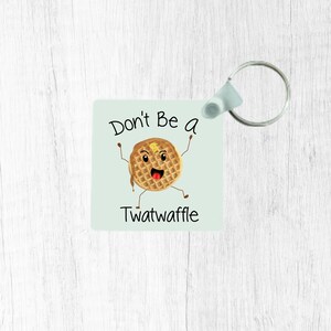 Peut inclure: Un porte-clés bleu clair avec une gaufre de dessin animé avec un visage et le texte "Don't Be A Twatwaffle".