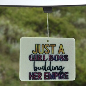 Pu&ograve; includere: Profumatore per auto bianco con il testo "Just a Girl Boss building her empire" in lettere colorate.