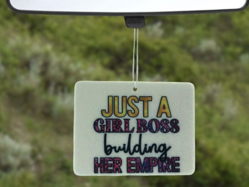 Pu&ograve; includere: Profumatore per auto bianco con il testo "Just a Girl Boss building her empire" in lettere colorate.