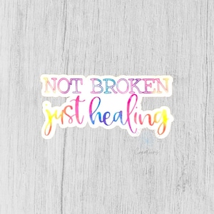 以下が含まれることがあります： 虹色のグラデーションで「NOT BROKEN just healing」というテキストが書かれたカラフルなステッカー。ステッカーは白い背景の上にあります。
