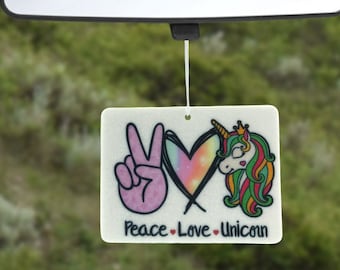 Può includere: Profumatore per auto bianco con un segno di pace rosa, un cuore arcobaleno e un unicorno verde, rosso e giallo con una corona. Il testo recita "Peace Love Unicorn".