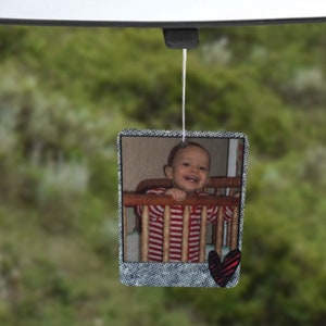 May include: A car air freshener featuring a photograph of a young child in a cot. The photo is set in a denim-style frame with a red heart.