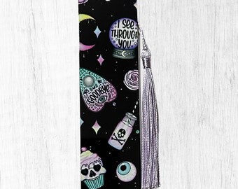 May include: A black bookmark with a variety of spooky illustrations, including a crystal ball, Ouija board, skull cupcake, and a bottle with a skull and crossbones. The text "I SEE THROUGH YOU" is visible. A silver tassel hangs from the top.