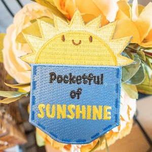 May include: A blue denim patch with yellow stitching and the text "Pocketful of Sunshine" in black. A yellow embroidered sun with a smiling face is above the text.