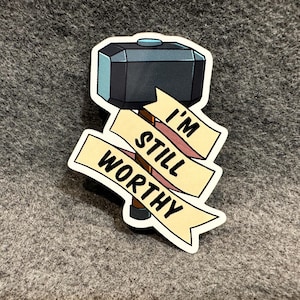 May include: A sticker featuring a cartoon hammer with a banner that reads "I'm Still Worthy". The hammer is black with a blue handle and the banner is yellow with brown text.