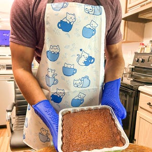 May include: A white apron with a blue and white pattern of cats in teacups. The apron is being worn by a person wearing a purple shirt. The person is holding a baking dish with a brown baked good inside. The person is wearing blue oven mitts.