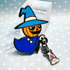 Puede incluir: Un pin de esmalte con temática de Halloween que presenta una figura con cabeza de calabaza que lleva un sombrero de bruja y una capa azules. El pin tiene un contorno negro e incluye un dije de linterna colgante. La calabaza es naranja con una cara negra tallada.
