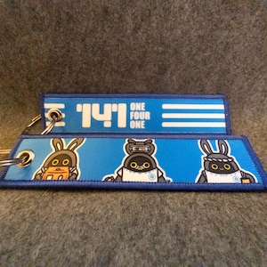 May include: Two blue keychains with cartoon robot-like characters and the text "141 ONE FOUR ONE". The keychains have a blue background with white and black details. Each keychain has a metal ring.
