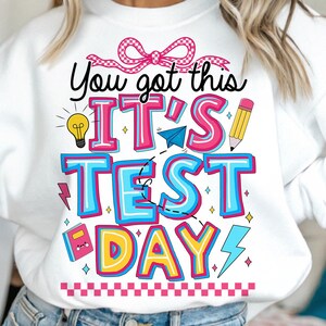 May include: White sweatshirt with a colorful graphic that says "You got this IT'S TEST DAY!" with a bow, lightbulb, airplane, pencil, and other fun illustrations.