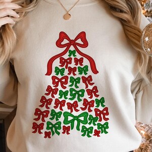 May include: Cream-colored sweatshirt featuring a Christmas tree design made of red and green bows. The bows are arranged in a tree shape, with a large red bow at the top and a mix of red and green bows forming the tree's body. A festive holiday apparel item.