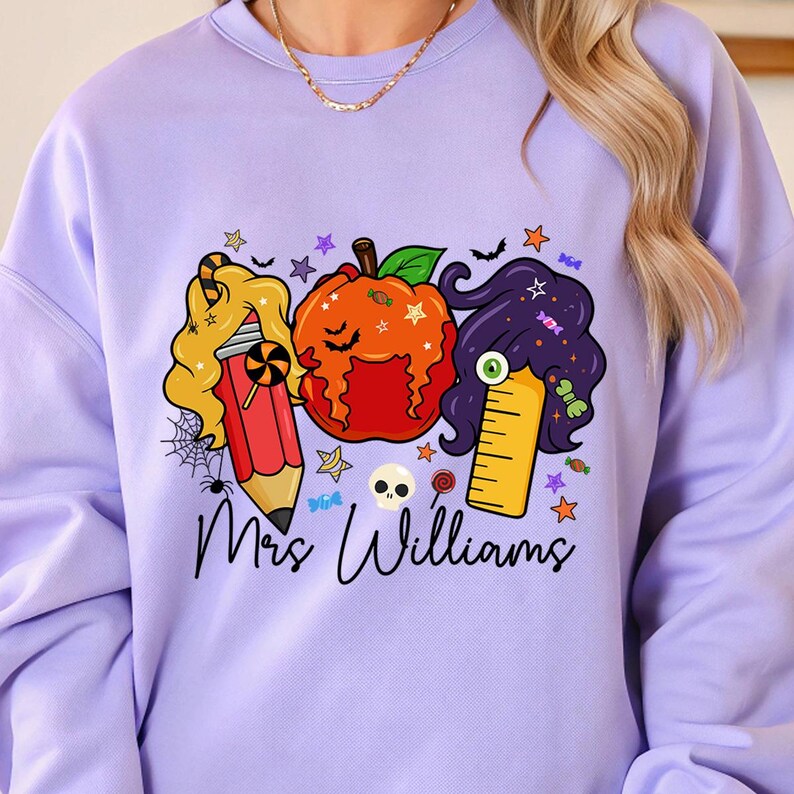 May include: A light purple sweatshirt featuring a Halloween-themed design. The design includes a pencil, pumpkin, and ruler with candy and star accents. The name "Mrs. Williams" is written in cursive below the graphics.