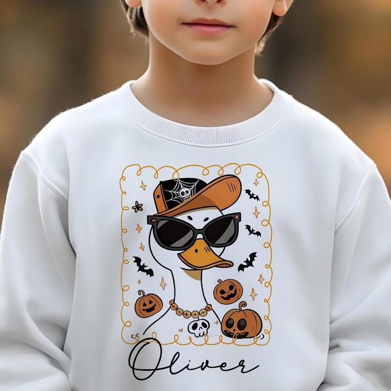 Op de afbeelding: Witte sweatshirt met een Halloween-thema eend met een zonnebril en een hoed. Het ontwerp bevat pompoenen, vleermuizen en de naam "Oliver". De eend draagt een gouden ketting met een schedel hanger.