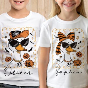 Op de afbeelding: Witte T-shirts met Halloween-eendenontwerpen. E&eacute;n eend draagt een zonnebril, een pet en een ketting, met de naam "Oliver" eronder. De andere eend heeft een strik, een zonnebril en een ketting, met de naam "Sophia".