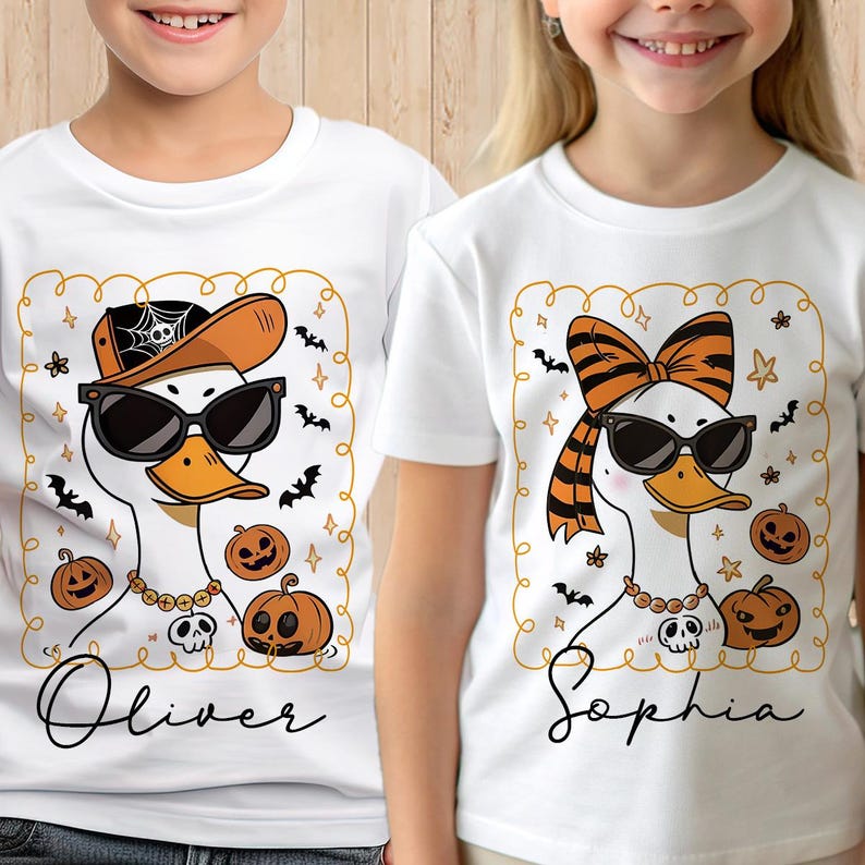Op de afbeelding: Witte T-shirts met Halloween-eendenontwerpen. E&eacute;n eend draagt een zonnebril, een pet en een ketting, met de naam "Oliver" eronder. De andere eend heeft een strik, een zonnebril en een ketting, met de naam "Sophia".