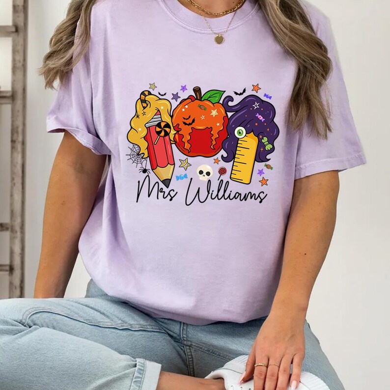 May include: A light purple t-shirt featuring a Halloween-themed graphic. The graphic includes a pencil, pumpkin, and ruler with hair, stars, and the text "Mrs Williams". The shirt is made of a soft material.