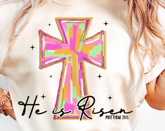 Op de afbeelding: Wit T-shirt met een kleurrijk abstract kruisontwerp en de tekst "He is Risen" met de bijbelverwijzing "Mattheüs 28:6".
