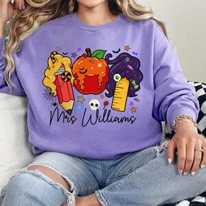 May include: A lavender sweatshirt featuring a Halloween-themed graphic. The design includes a pumpkin, pencil, and ruler with cartoon hair, candy, and the text "Mrs. Williams". The sweatshirt is a comfortable, casual style.