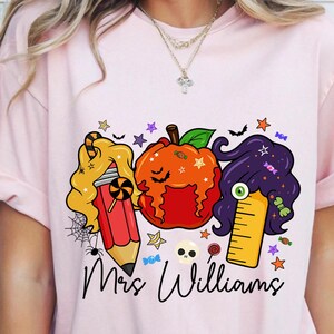 May include: Pink t-shirt featuring a Halloween-themed graphic. The design includes a pencil, pumpkin, and ruler with candy and star accents. The text "Mrs. Williams" is written in a script font below the graphic.