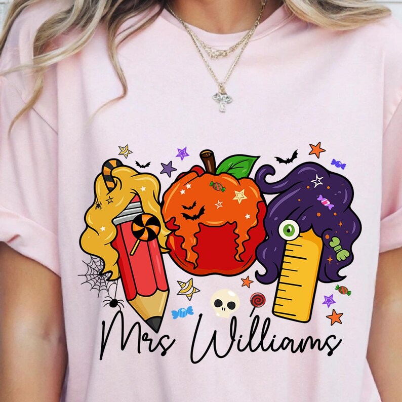 May include: Pink t-shirt featuring a Halloween-themed graphic. The design includes a pencil, pumpkin, and ruler with candy and star accents. The text "Mrs. Williams" is written in a script font below the graphic.