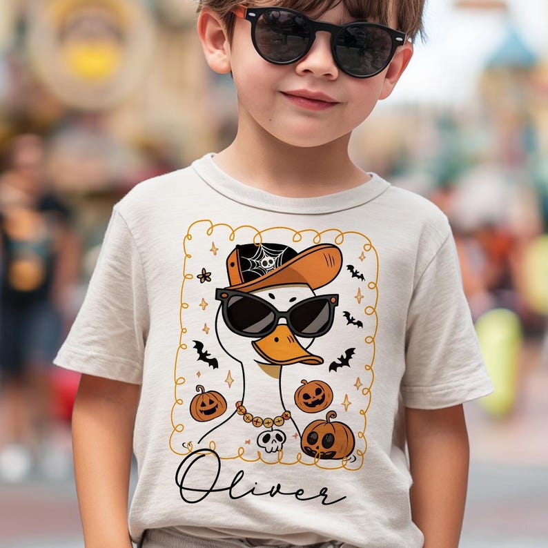 Op de afbeelding: Beige T-shirt met korte mouwen met een cartoon eend met een zonnebril, een hoed met een spinnenweb en een ketting met een schedel hanger. Het ontwerp bevat pompoenen en vleermuizen, met de naam "Oliver" in cursief.