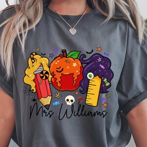 May include: Dark gray t-shirt featuring a Halloween-themed graphic. The design includes a pencil, pumpkin, and ruler with candy and star accents. The text "Mrs. Williams" is written in a script font below the graphic.