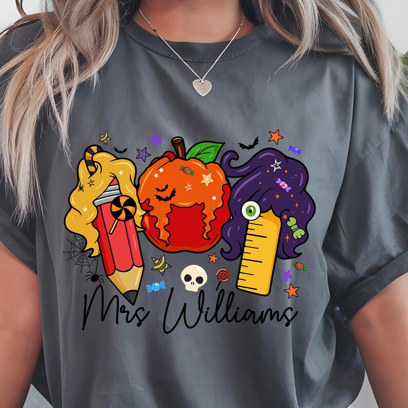 May include: Dark gray t-shirt featuring a Halloween-themed graphic. The design includes a pencil, pumpkin, and ruler with candy and star accents. The text "Mrs. Williams" is written in a script font below the graphic.