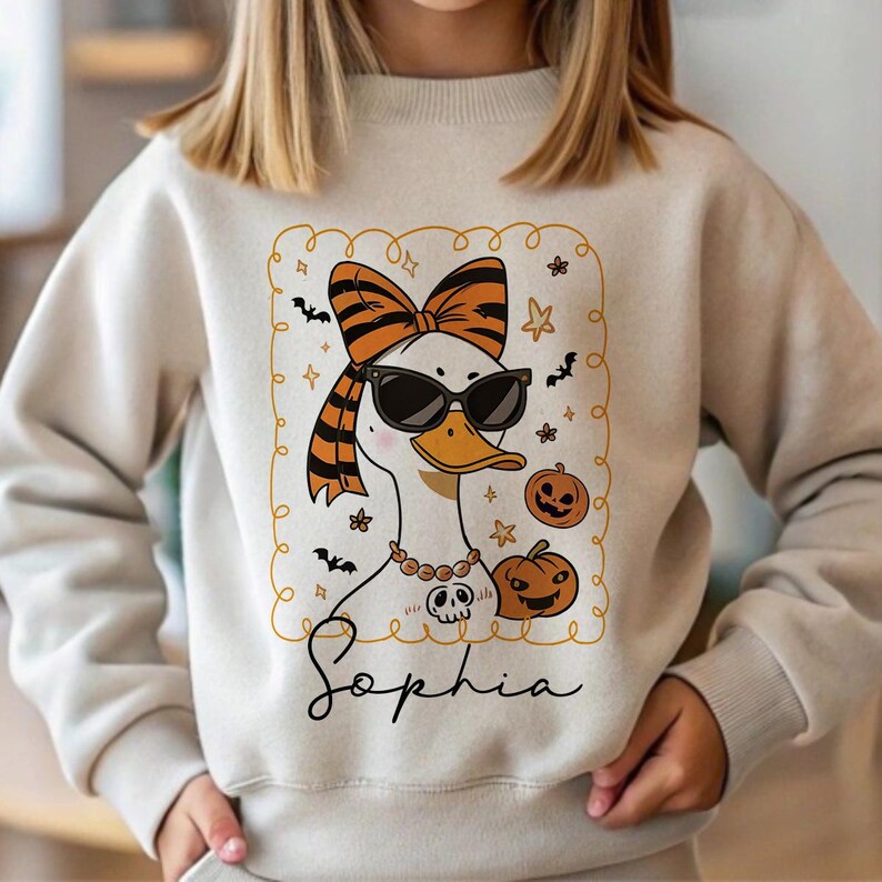 Op de afbeelding: Beige sweatshirt met een Halloween-thema. De afbeelding toont een witte eend met een zonnebril, een strik en een ketting met een schedel hanger. Pompoenen, vleermuizen, sterren en de naam "Sophia" zijn ook in het ontwerp opgenomen.