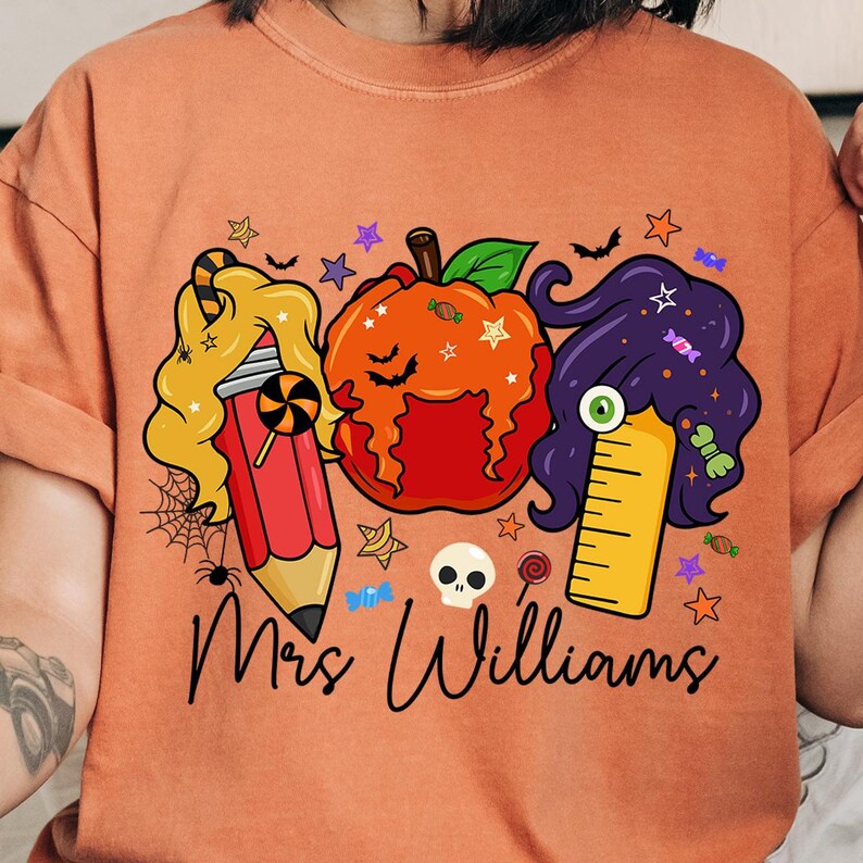 May include: Orange t-shirt featuring a Halloween-themed design. The graphic includes a pencil with blonde hair, a pumpkin, and a ruler with purple hair. The text "Mrs. Williams" is written in a script font.