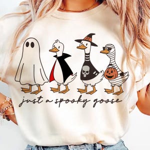 May include: Cream-colored t-shirt featuring a Halloween-themed design. The graphic depicts four cartoon geese dressed as a ghost, vampire, witch, and mummy. The text "just a spooky goose" is written in a script font below the geese.