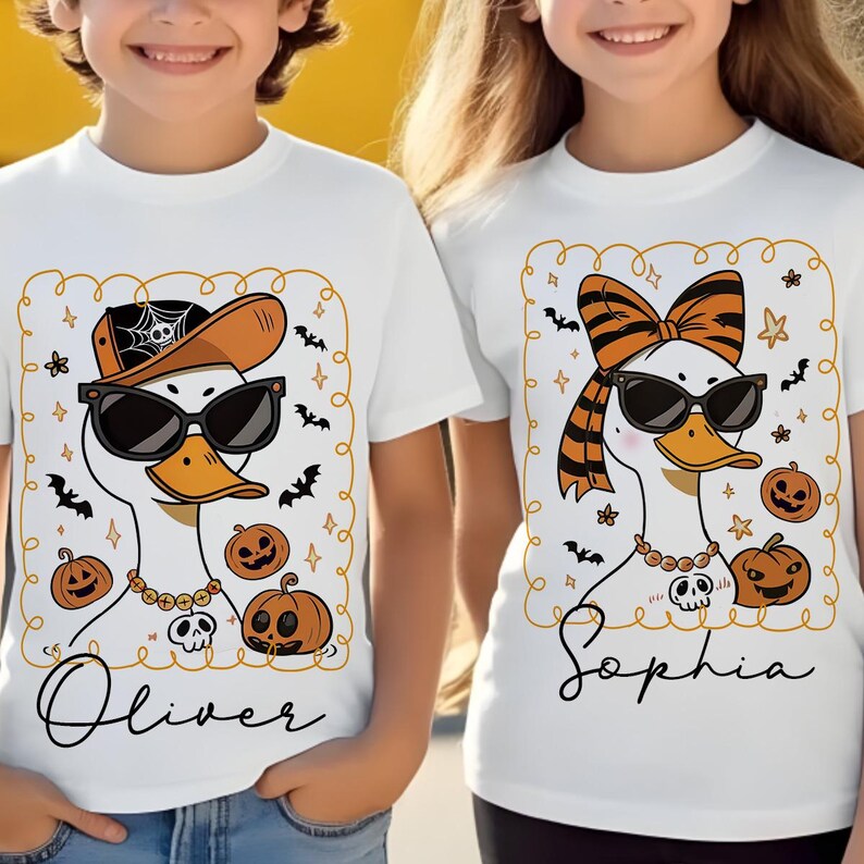 Op de afbeelding: Witte T-shirts met Halloween-eendengraphics. E&eacute;n eend draagt een pet en zonnebril, de andere een strik en zonnebril. Beide hebben kettingen, pompoenen en vleermuizen. De naam "Oliver" staat onder de ene, en "Sophia" onder de andere.